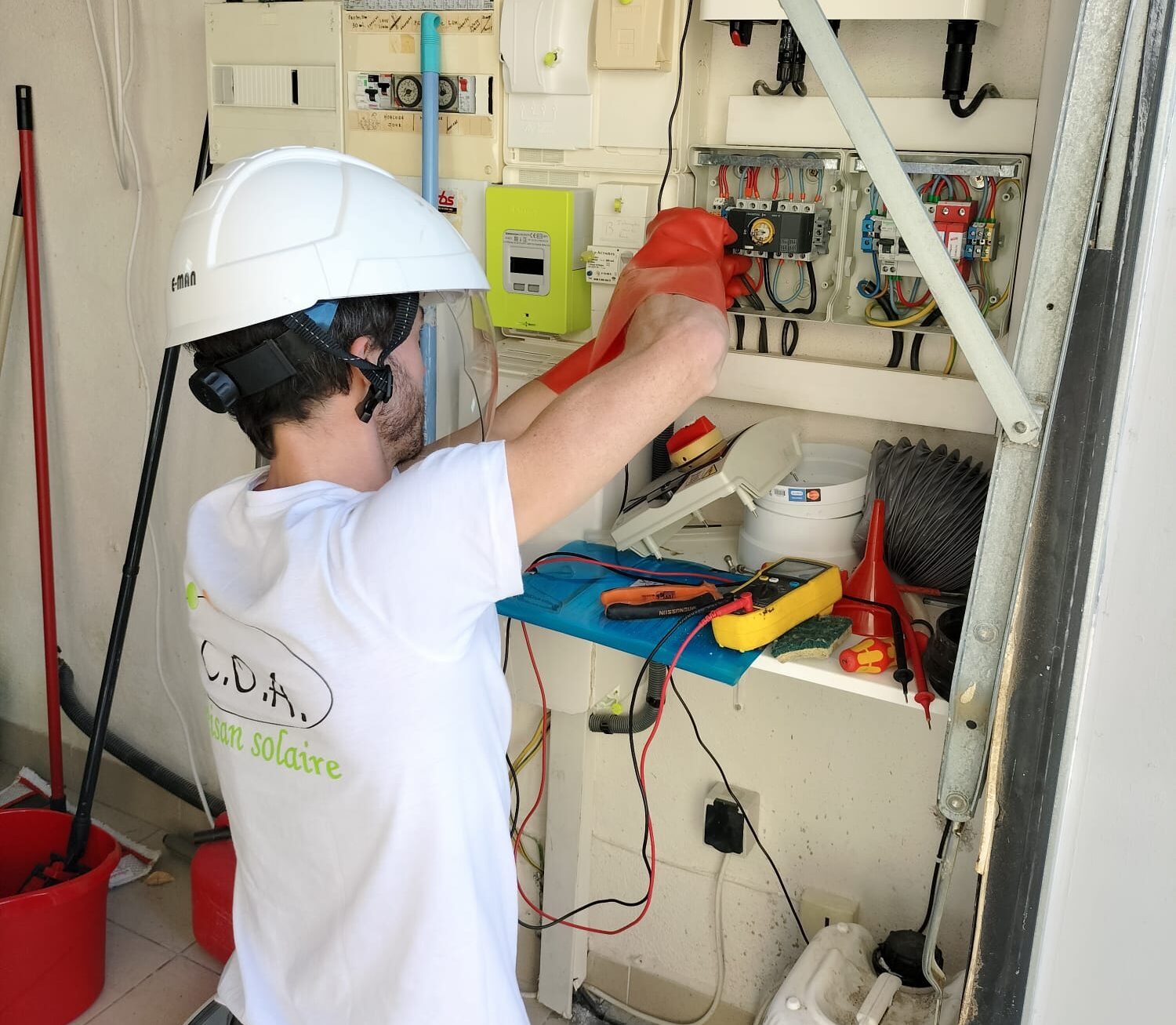 Technicien en casque et gants isolants mesure un tableau électrique photovoltaïque, multimètre et outils visibles.