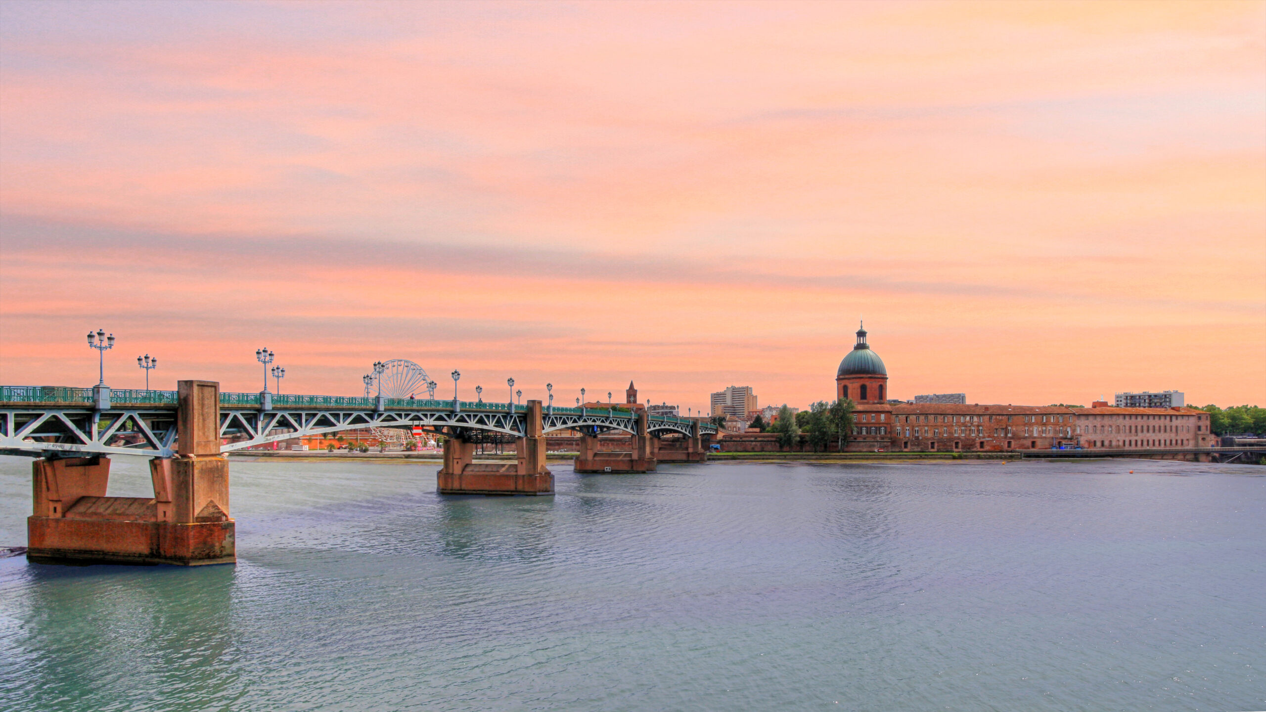 Panorama de la Garonne à Toulouse : au premier plan le Pont Saint-Pierre en structure métallique verte posé sur des piles de pierre, en arrière-plan la façade ocre et le dôme de l’Hôtel-Dieu Saint-Jacques, sous un ciel de fin de journée aux teintes rosées.