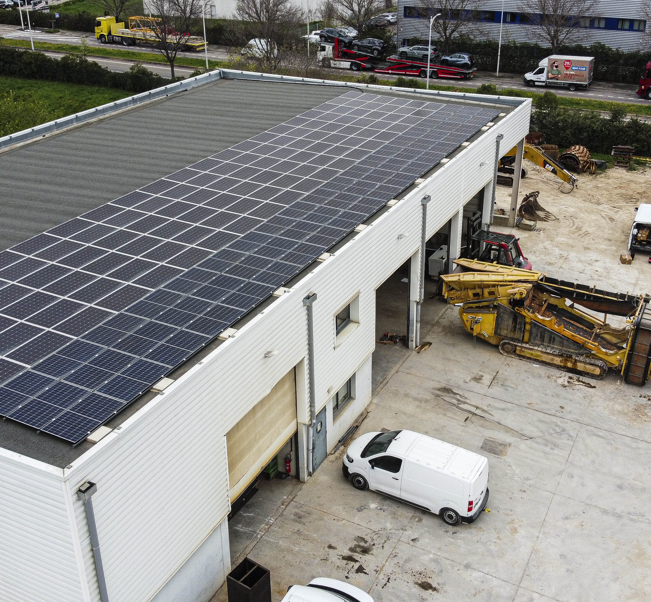Vue oblique d’un bâtiment industriel blanc de Crozel T.P, avec une rangée de panneaux solaires bleus fixés sur son toit plat et une aire de stockage d’engins et véhicules à proximité.