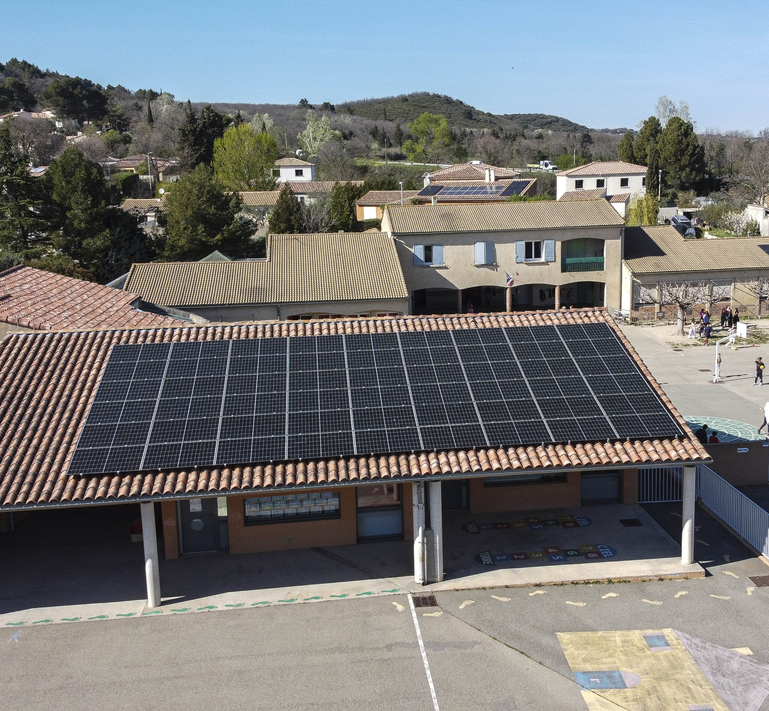 Vue en légère plongée d’un préau à toit en tuiles canal équipé de modules solaires bleus affleurants, avec la cour de récréation et les bâtiments de l’école en arrière-plan sous un ciel dégagé.
