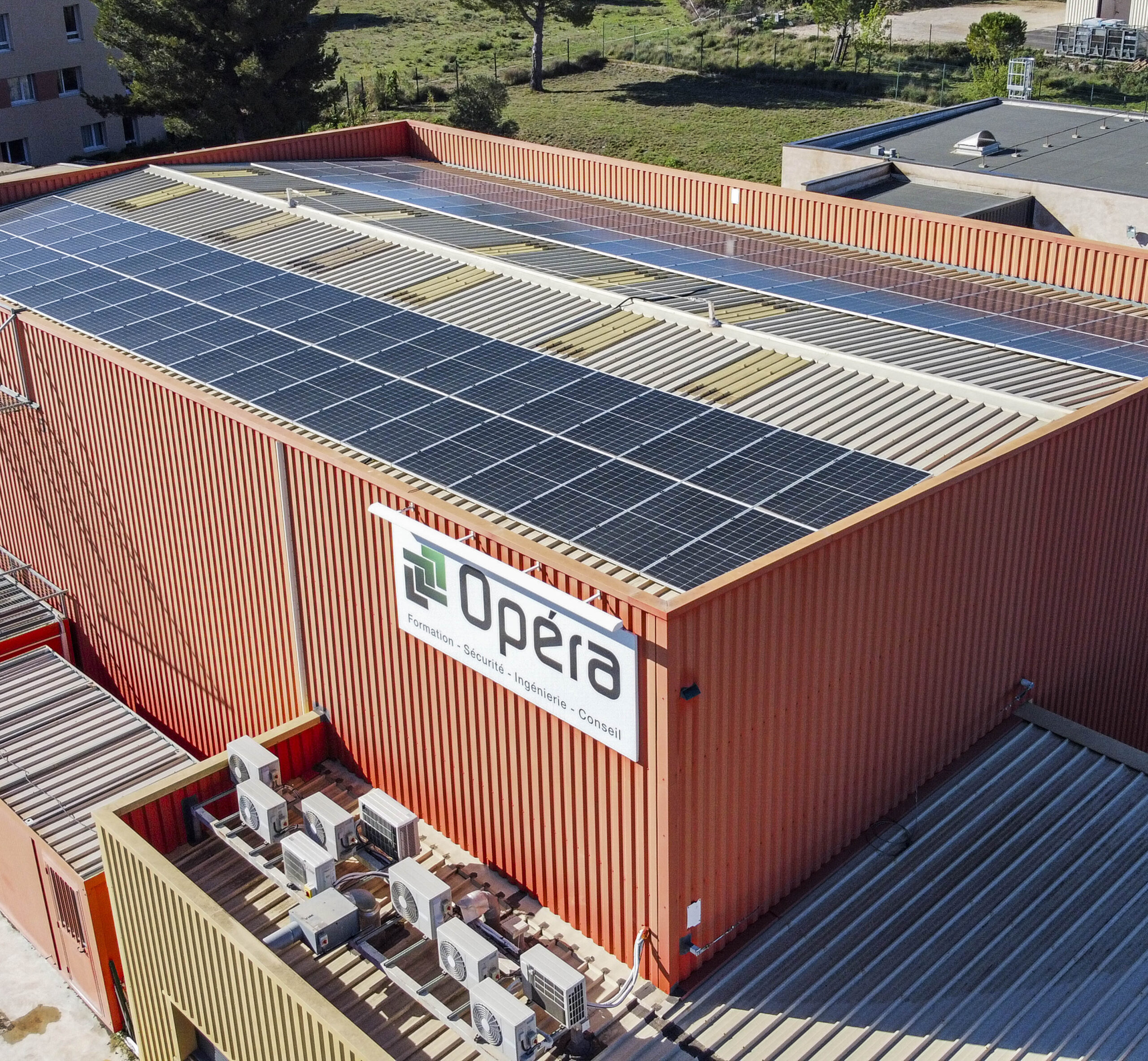 Vue en perspective aérienne d’un bâtiment rouge “Opéra Formation – Sécurité • Ingénierie • Conseil” avec des panneaux solaires alignés sur sa toiture bac acier.