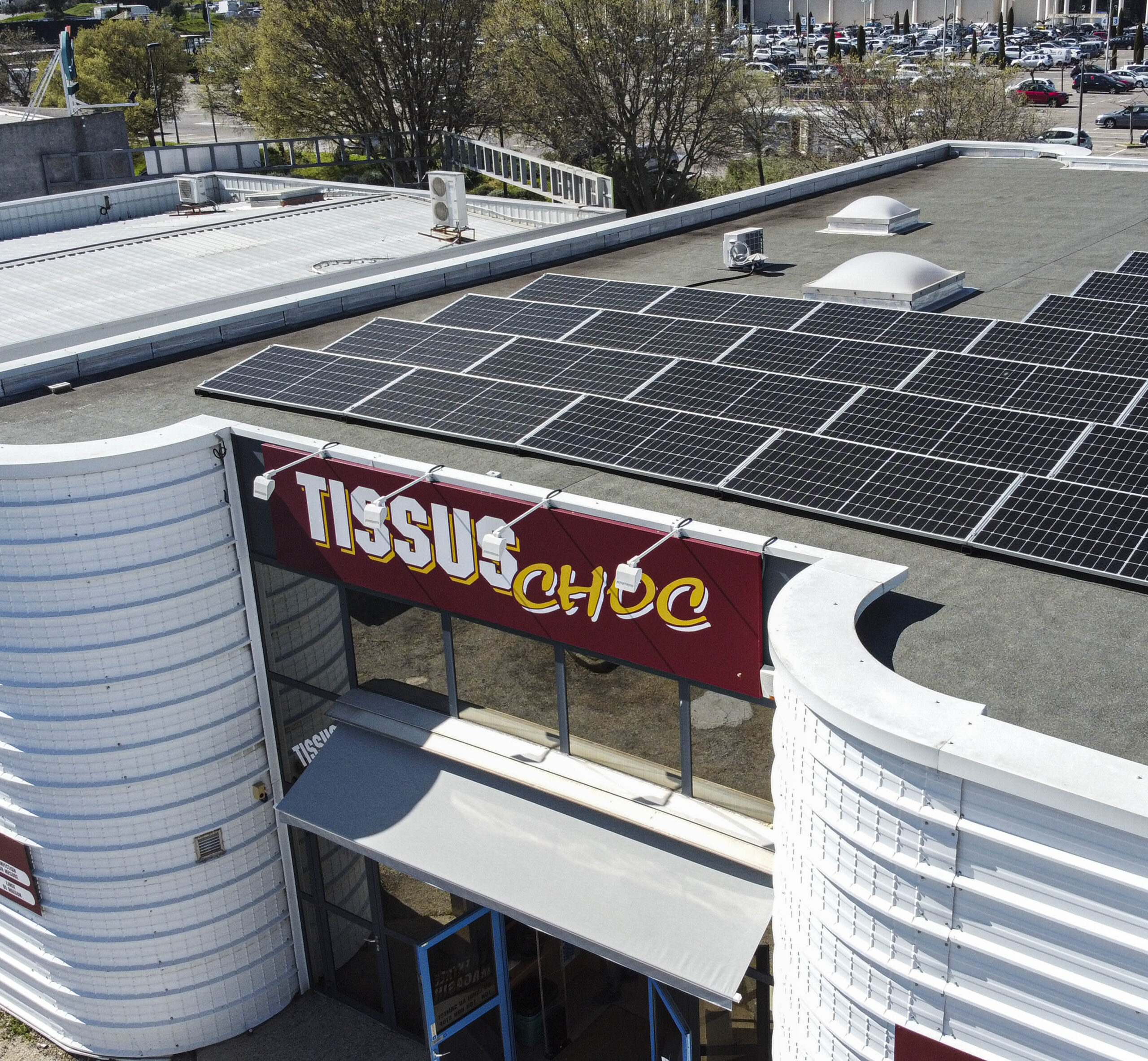 Vue en légère plongée sur un toit plat équipé de panneaux solaires au-dessus de la façade du magasin “Tissus Choc”, avec sa enseigne rouge et blanche et des baies vitrées sous un ciel dégagé.
