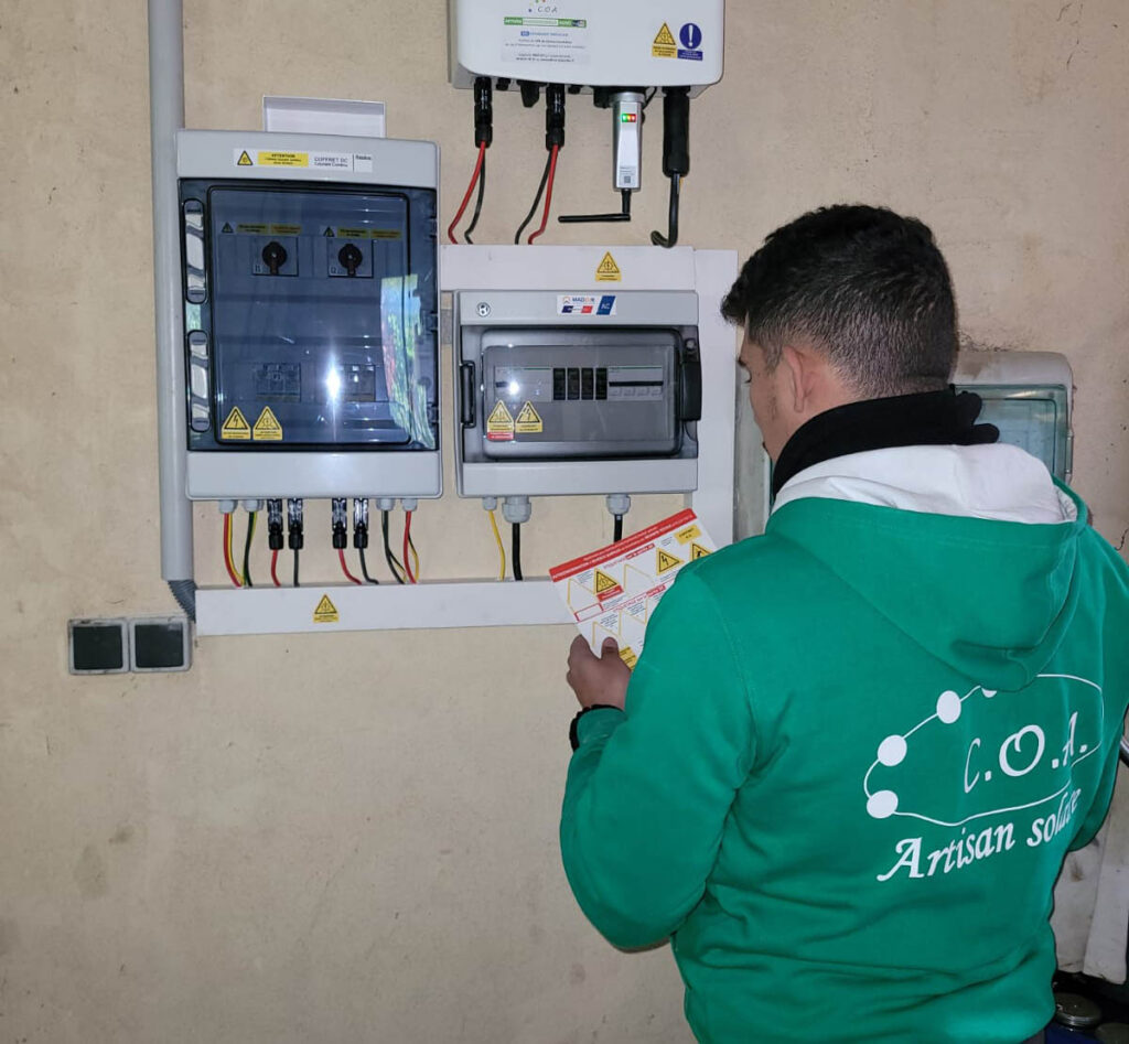 Un installateur portant un sweat C.O.A « Artisan solaire » consulte un schéma devant un coffret électrique DC et AC relié à un onduleur solaire.