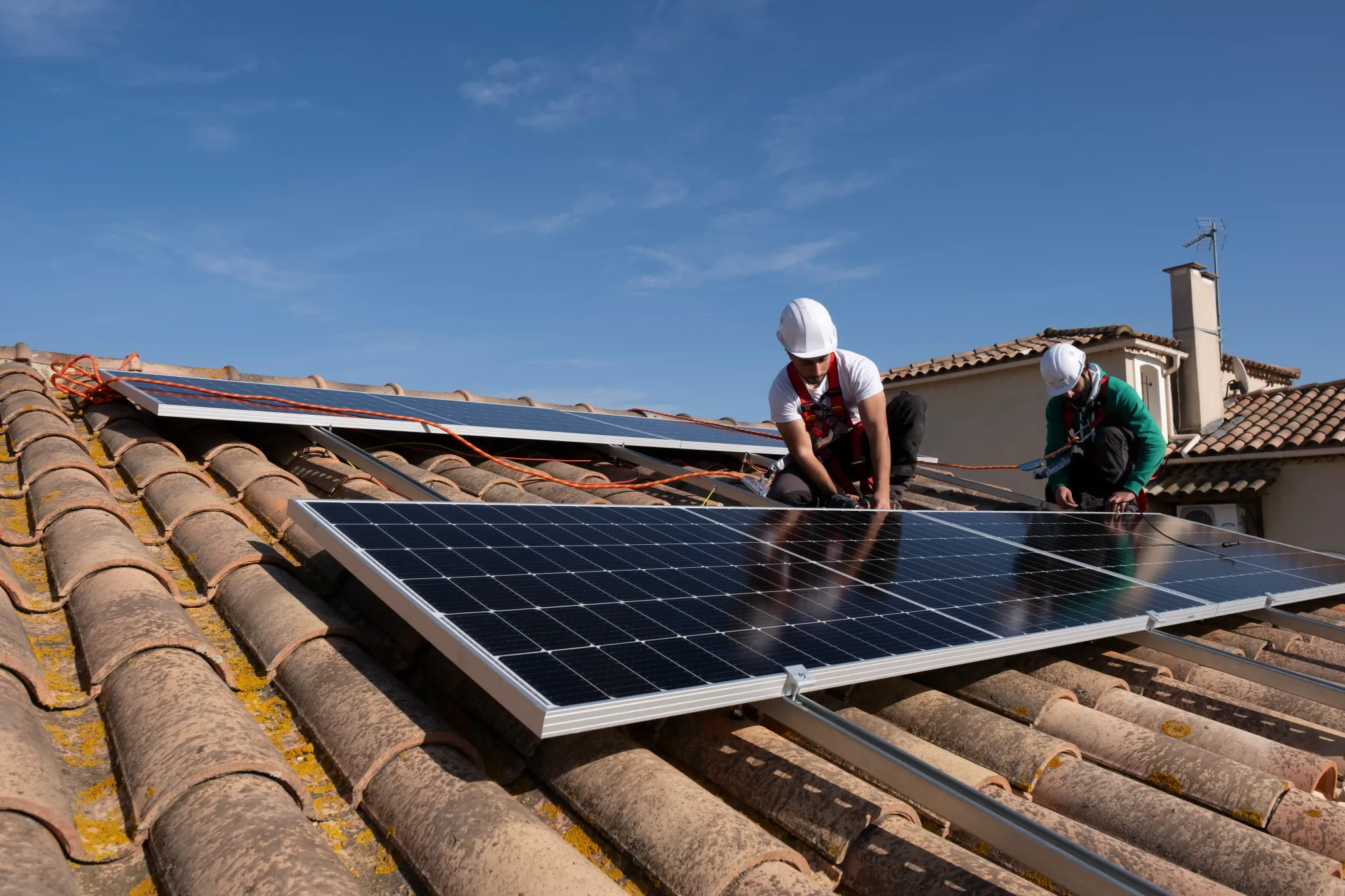Deux techniciens installent des panneaux photovoltaïques sur une toiture en tuiles, avec harnais de sécurité et câblage visible.