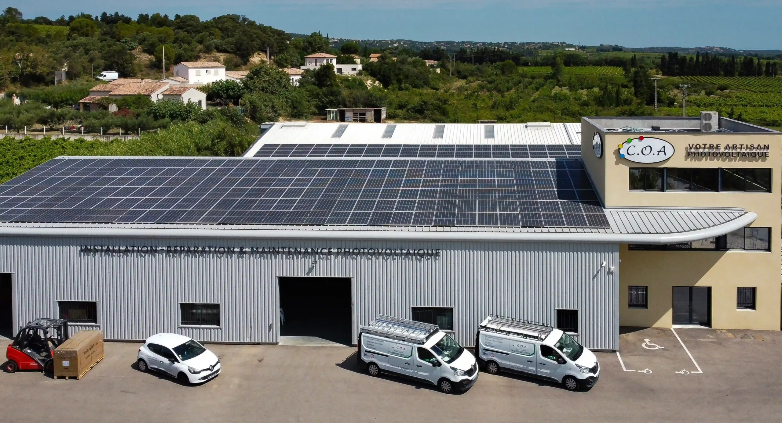Scène devant les locaux de C.O.A montrant plusieurs véhicules utilitaires de l’entreprise stationnés sous une vaste toiture photovoltaïque, avec un bâtiment industriel équipé de panneaux solaires et un paysage verdoyant en arrière-plan.