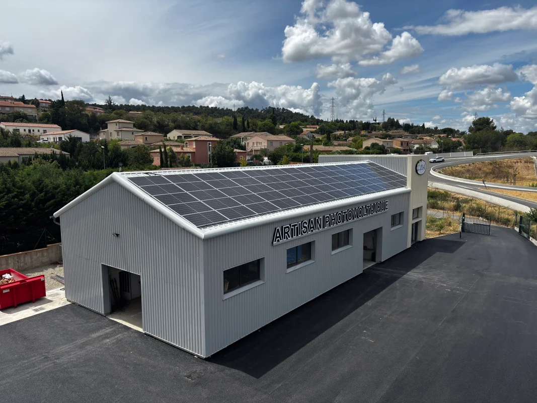 Photo prise en plongée d’un bâtiment métallique gris avec des panneaux solaires sur le toit et le sigle “ARTISAN PHOTOVOLTAÏQUE” sur sa façade, entouré d’un parking noir bitumé, d’une haie de cyprès et d’une route en contrebas.